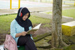 © Fajar - Serene young woman in a hijab and red headphones, deeply engrossed in reading a book while studying outdoors on a tranquil park bench, fostering knowledge and personal growth