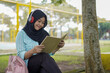 © Fajar - Serene young woman in a hijab and red headphones, deeply engrossed in reading a book while studying outdoors on a tranquil park bench, fostering knowledge and personal growth