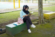 © Fajar - Serene young woman in a hijab and red headphones, deeply engrossed in reading a book while studying outdoors on a tranquil park bench, fostering knowledge and personal growth
