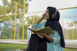 © Fajar - Serene young woman in a hijab and red headphones, deeply engrossed in reading a book while studying outdoors on a tranquil park bench, fostering knowledge and personal growth
