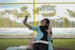 © Fajar - Young woman in hijab and headphones using a smartphone to record a video outdoors with a notebook and pen nearby, capturing a moment of content creation and learning in a casual setting