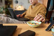 © Mediaphotos - Man sitting at desk using laptop while reaching for bowl of colorful candy, focus on hand and snack, blurred background suggesting home or office setting