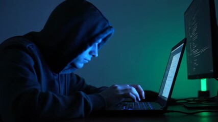 Hooded male hacker typing on a laptop with glowing screen in a dark room with an additional monitor, cybersecurity concept footage.