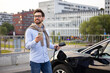 © sofiko14 - A man smiles while holding a phone and charging his electric car in an urban setting.
