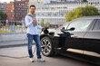 © sofiko14 - A man smiles while charging his electric vehicle with a mobile phone in hand, showcasing modern technology and sustainable transportation.