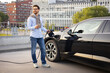 © sofiko14 - A man smiles while charging his electric vehicle at a charging station, holding his phone.