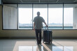 © samael334 - Man traveler standing with suitcase looking out airport window at runway at airport terminal. Male tourist with luggage waiting for flight departure near boarding area. Travel, vacation