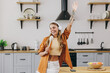 © ViDi Studio - Young housewife woman wear orange shirt casual clothes listen to music in headphones use wooden spoon pov microphone sing sit at table in light kitchen at home alone. Lifestyle cooking food concept.