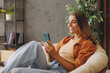 © ViDi Studio - Side view young woman wear orange casual clothes hold in hand use mobile cell phone sits in chair stay at home hotel flat rest relax spend free spare time in living room indoor. People lounge concept.