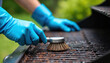 © miss irine - Person wearing blue gloves cleans barbecue grill with brush. Hand holds brush, scrub grill surface. Plants in background. Outdoor cooking preparation. Cleaning grill with tool.