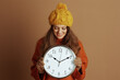 © Alliance - A smiling woman in a yellow beanie and orange turtleneck sweater holds a large wall clock, looking down at it. Her warm, cheerful expression against a brown background evokes a cozy autumn mood.