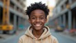 © Vadym - African American teenage boy smiles outdoors in beige hoodie. He has curly dark hair and a happy expression. Blurred city street background with construction elements.