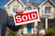 © Vitalii Shkurko - A person in a suit proudly holds a sold sign in front of a beautiful house, showcasing a successful real estate transaction on a clear day with blue skies