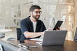 © muse studio - Man working on digital tablet and laptop in modern office setting while engaged in a productive meeting with colleagues