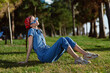 © SHOTPRIME STUDIO - girl relaxing on grass wearing blue denim jumpsuit and red bandana enjoying sunny day outdoors with green trees background and casual sneakers