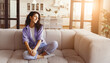 © Prostock-studio - A young woman sits comfortably on a soft beige sofa, smiling as sunlight streams through large windows. The bright and airy living room features modern decor and a cozy atmosphere.