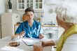 © shurkin_son - Side view of essential in-home medical support and companionship for elderly, caring nurse in blue uniform, with stethoscope on shoulders conducting health assessment for senior woman at kitchen table