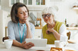 © shurkin_son - Female volunteer of retirement home nursing her senior female patient, watching series together or grandmother videochatting with her children using tablet sitting at kitchen table during breakfast