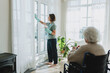 © shurkin_son - Cleaning service for seniors concept. Female cleaner in gloves washing big panoramic windows in cozy living-room using detergent and talking to old disabled lady home owner in wheelchair