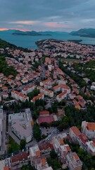Wall Mural - Aerial drone flies over Dubrovnik town, capturing red rooftops, houses, and surrounding mountains glowing warmly in golden light during a calm autumn sunset across the scenic Croatian coastline.