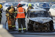 © Seventyfour - Group of firefighters and emergency responders extinguishing burning car on city street, wearing protective gear and helmets, smoke rising from damaged vehicle, traffic visible in background