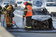 © Seventyfour - Group of firefighters extinguishing burning car on busy city street, wearing protective uniforms and helmets, smoke rising from damaged vehicle, traffic visible in background