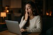 © Loginova - Tired woman yawning while working late at night on laptop in cozy dimly lit room.