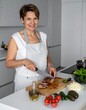 © Ant - A woman in an apron slices meat on a wooden cutting board surrounded by fresh vegetables and olive oil in a modern kitchen.
