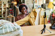 © pressmaster - Young adult Black woman smiling while presenting jacket to smartphone camera, standing behind counter in thrift shop, engaging in online selling or live streaming clothing items