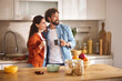 © Stockphotodirectors - A cheerful couple stands side by side in a sleek kitchen, holding coffee mugs and smiling. Sunlight streams in, enhancing a cozy morning atmosphere filled with vibrant colors.