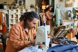 © pressmaster - Caucasian young adult man using sewing machine while working on clothing repair at thrift shop, Black young adult woman browsing clothing racks in background, shelves filled with garments visible