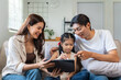 © amnaj - Asian parents and young daughter sitting on sofa, smiling and using a digital tablet for online learning, bonding, and entertainment