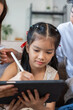 © amnaj - Little Asian girl learning on device, using a stylus to draw while her parents are supervising and supporting her