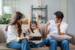 © amnaj - Young Asian family sitting on sofa, parents watching happy daughter raising arms while winning on tablet in living room