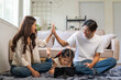 © amnaj - Happy family bonding at home, parents high five while their daughter uses a tablet on the living room floor, cheerful connection