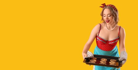 Wall Mural - Young pin-up housewife holding baking tray with cookies on yellow background with space for text