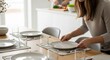 © Таня Андреева - A woman setting a wooden dining table with plates, glasses, and placemats. Focus on home lifestyle, meal preparation, and modern domestic life in a bright space.