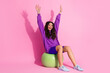 © deagreez - Joyful young woman in vibrant activewear sitting on exercise ball against a pink background, raising hands in excitement