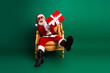 © deagreez - Santa Claus sitting on a golden chair holding a red gift, wearing sunglasses, in front of a vibrant green backdrop.