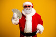 © deagreez - Santa Claus in vibrant costume holding money and wearing sunglass, posing with a cheerful smile against a bright yellow background