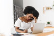 © etonastenka - African American woman sitting at home table with laptop, papers, and calculator, looking stressed and upset. Concept of financial problems, remote work challenges, budgeting, and personal finance