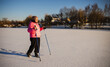 © StockMediaSeller - Elderly woman walking with trekking poles on a frozen snow-covered field during sunny winter morning. She wears a pink jacket and looks happy and healthy while exercising outdoors.