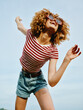 © SHOTPRIME STUDIO - Bright joyful portrait of a young woman with curly hair, sunglasses, striped tee, and denim shorts, jumping with carefree energy against a clear blue sky outdoors.