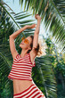 © SHOTPRIME STUDIO - A joyful woman wearing a red and white striped outfit dances with arms raised under sunny tropical palms, exuding carefree summer vibes and vibrant energy.
