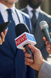 © pressmaster - Caucasian middle aged man wearing suit speaking to journalists holding microphones during press conference outdoors, hands of reporters visible in foreground, focus on media interaction