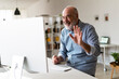 © BusinessClass by W61 - Cheerful senior businessman waving hand on video call through desktop computer in home office