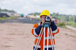 © kamonrat - A survey engineer wearing an orange reflective jacket and yellow helmet operates a leveling instrument on a tripod at a construction site