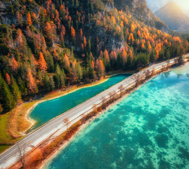 Wall Mural - Aerial view of scenic road running along turquoise alpine lake with clear water, surrounded by colorful autumn forest and rocky mountains. Beautiful alpine landscape in fall season, travel background