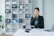 © MINAE - Young Asian businesswoman sitting at an office desk, holding a pen to her chin and carefully considering financial charts and reports while strategizing for future business growth