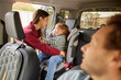 © Stockphotodirectors - A woman securely fastens a young child in a car seat while a man looks on from the driver's seat, showing a moment of connection and care during a family outing.